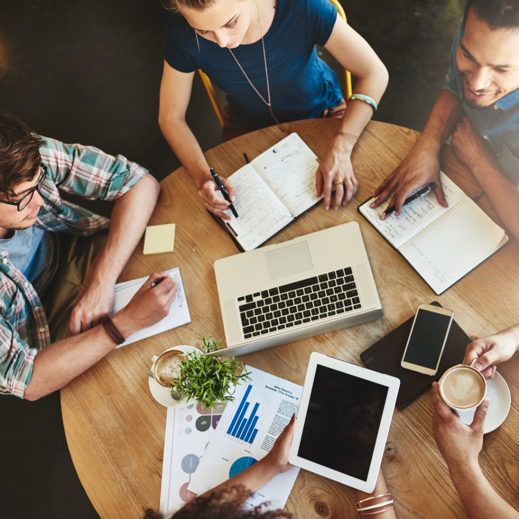 A group gathers around a round table with a laptop, notebooks, phones, and cups of coffee.
