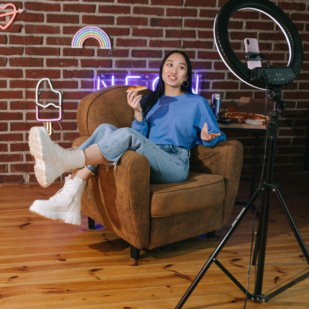 A girl sits on a brown couch, eating pizza while engaging with a live audience.